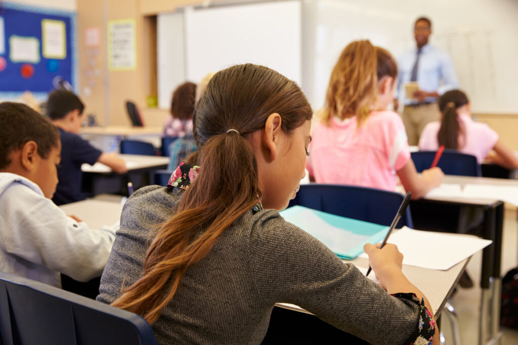 A classroom scene showing several students focused on their work. In the foreground, a girl with long hair is writing in a notebook, while a boy in a blue shirt is visible to the left. In the background, a teacher stands in front of a whiteboard, addressing the class, while other students sit at their desks, writing or looking ahead.