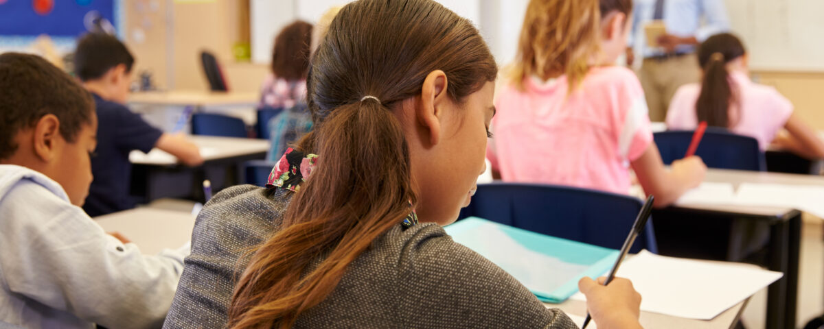 A classroom scene showing several students focused on their work. In the foreground, a girl with long hair is writing in a notebook, while a boy in a blue shirt is visible to the left. In the background, a teacher stands in front of a whiteboard, addressing the class, while other students sit at their desks, writing or looking ahead.