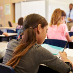 A classroom scene showing several students focused on their work. In the foreground, a girl with long hair is writing in a notebook, while a boy in a blue shirt is visible to the left. In the background, a teacher stands in front of a whiteboard, addressing the class, while other students sit at their desks, writing or looking ahead.