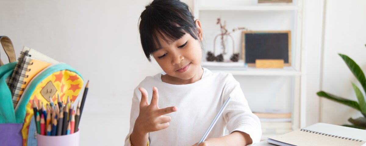 A young multilingual learner is seated at a table, focused on writing in a notebook while making a hand gesture. She has a look of concentration, surrounded by school supplies including colored pencils, paint, and sticky notes. In the background, shelves with decorative items and books can be seen, creating a cozy study environment.