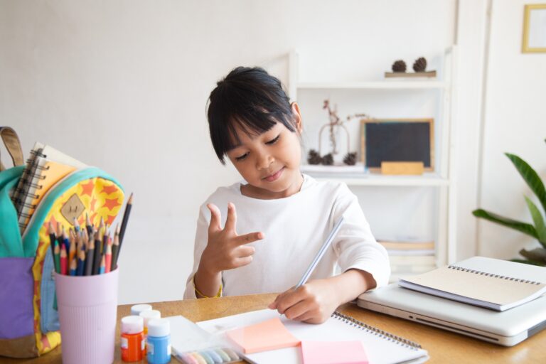 A young multilingual learner is seated at a table, focused on writing in a notebook while making a hand gesture. She has a look of concentration, surrounded by school supplies including colored pencils, paint, and sticky notes. In the background, shelves with decorative items and books can be seen, creating a cozy study environment.