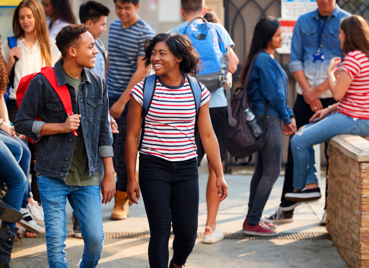 A group of high school students socializing outside on campus. Two students, one wearing a red-striped shirt and the other with a red backpack, walk together smiling. Other students in the background are chatting and sitting casually, creating a lively and friendly school atmosphere.