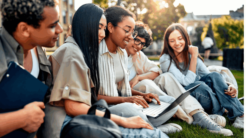 A group of five college students sitting together on the grass outdoors, smiling and looking at a laptop screen. They appear to be students collaborating or studying, with notebooks and backpacks nearby, in a sunny, relaxed campus-like setting.