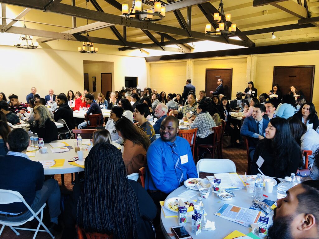 A large group of professionally dressed people seated at round tables in a spacious, well-lit room with exposed wooden beams and chandeliers, engaged in conversation and eating during a formal event.