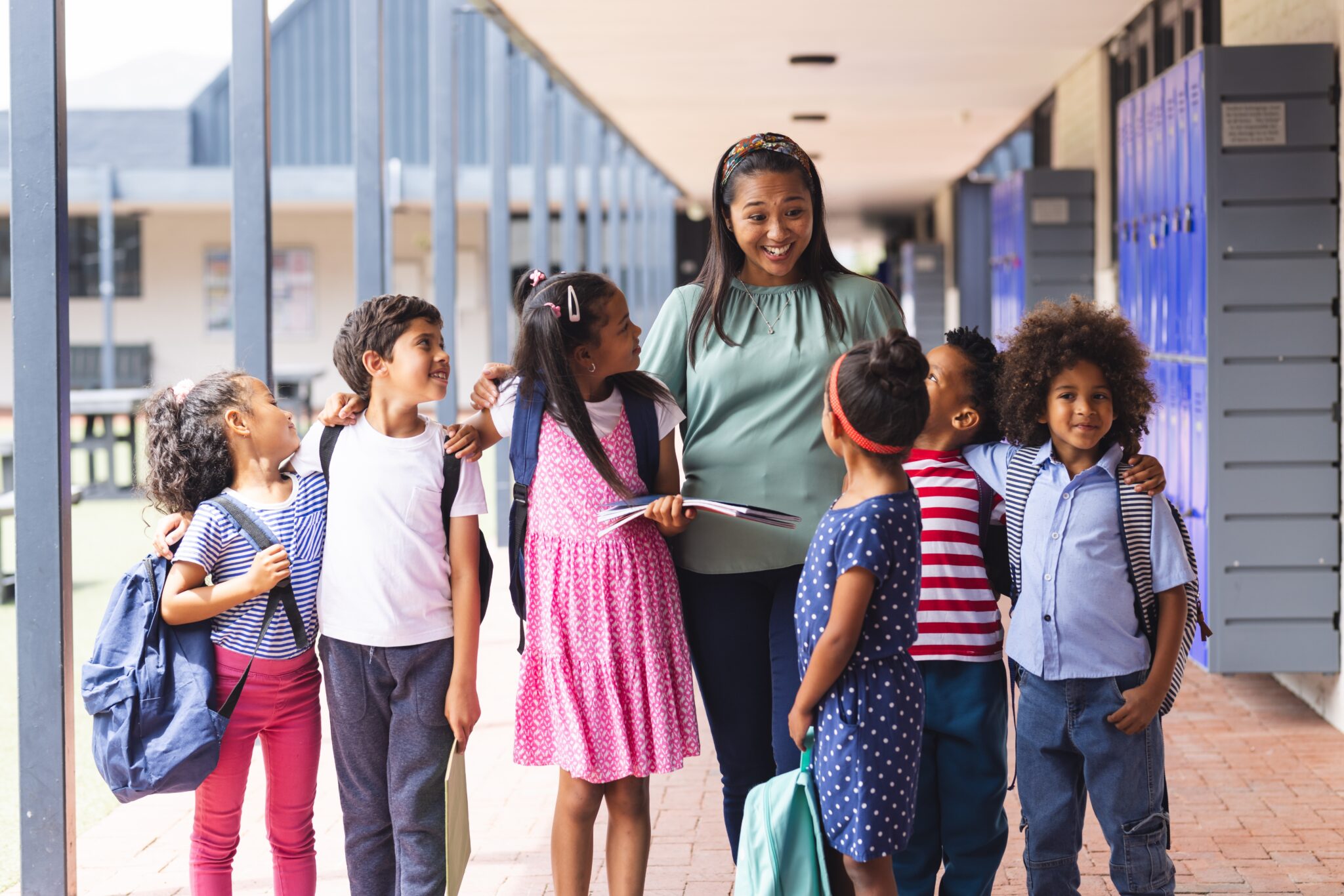 A teacher stands with a group of young students in a school hallway, with lockers on one side and an outdoor area visible on the other. The children are gathered closely around the teacher, some holding backpacks and books.