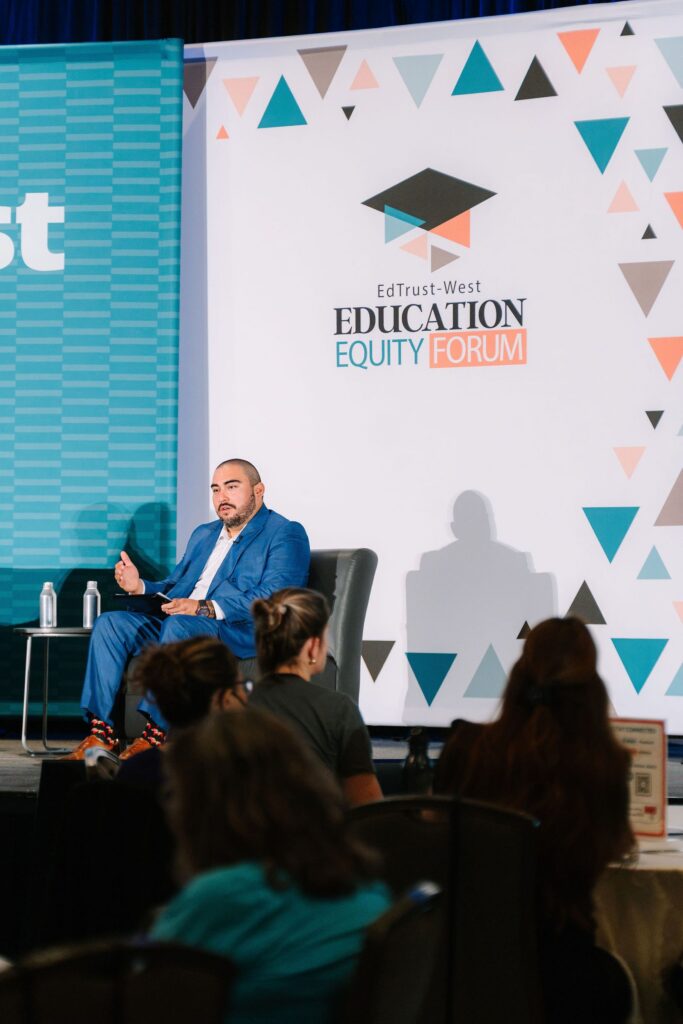 A speaker seated on stage addressing the audience in front of the EdTrust-West Education Equity Forum backdrop