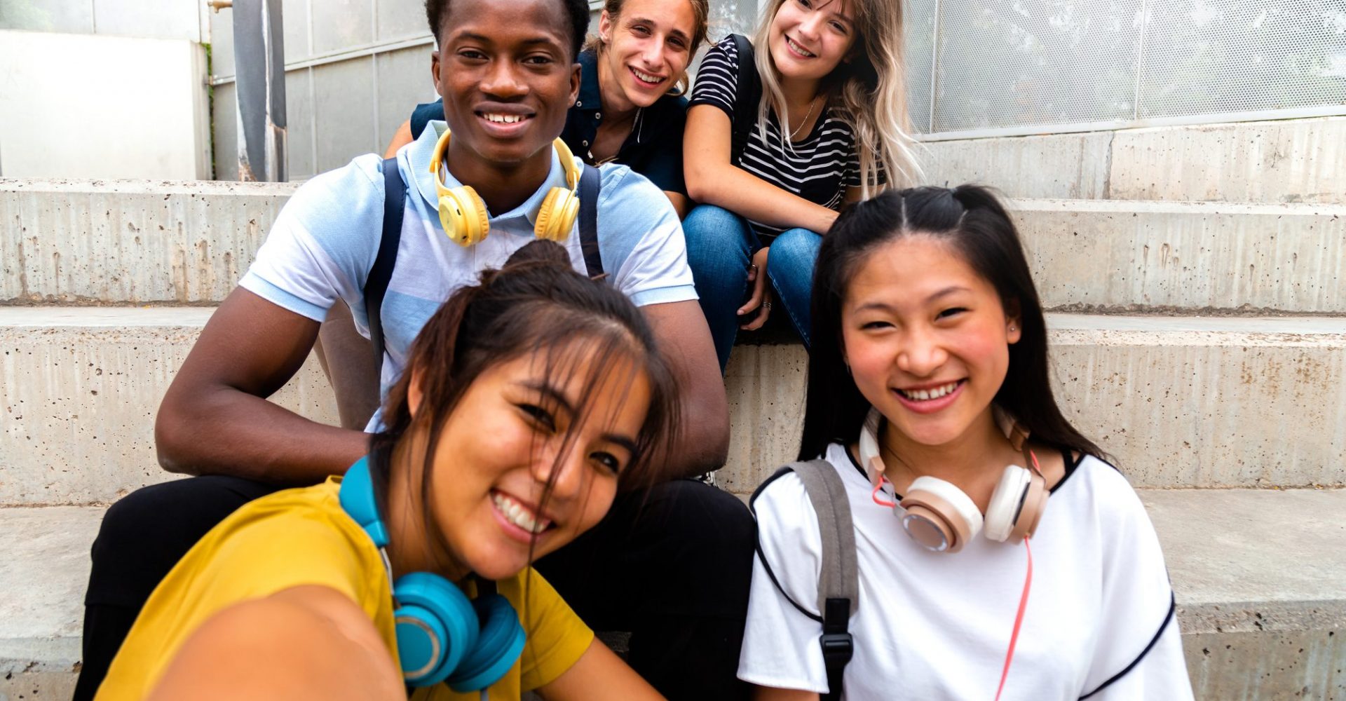 Happy smiling teen multiracial group of students looking at camera take selfie sitting on steps outside. Education and social media concepts.
